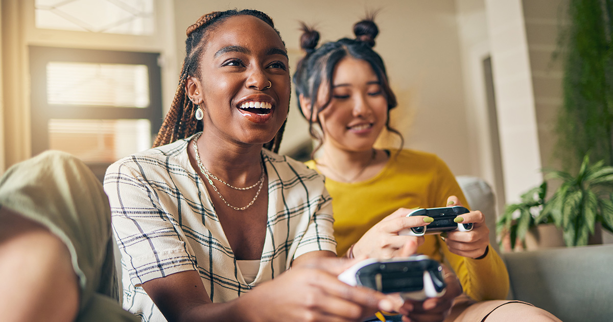 Two young women sit on a couch smiling while one games and the other is on her phone.